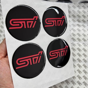 May include: A set of four circular, black, domed stickers featuring the red STI logo. Each sticker has a glossy finish and is arranged on a white backing sheet with a repeating 3M logo pattern, designed for application as automotive emblems.