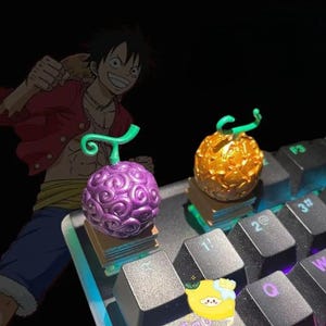 Puede incluir: Dos detalladas Frutas del Diablo de resina, una morada y otra dorada, se asientan sobre pequeñas pilas de libros en un teclado. Un personaje de dibujos animados está en el fondo. La fruta morada tiene un patrón arremolinado, y la fruta dorada tiene una superficie texturizada.