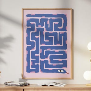 Può includere: Una stampa incorniciata con un disegno a labirinto blu e una testa di serpente su sfondo rosa. È appesa sopra un mobile in legno chiaro, con una lampada dorata e una scultura decorativa. Il mobile è largo 150 cm.
