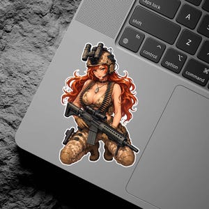 May include: A sticker featuring an anime-style illustration of a female soldier kneeling, holding a rifle. She wears a camouflage uniform, a helmet with night vision goggles, and has long red hair. The sticker is placed on a laptop.