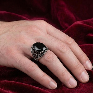 May include: A silver ring with a large, oval-cut black gemstone. The ring has an ornate, detailed design around the stone, with smaller black accents. The ring is displayed on a hand against a dark red background.