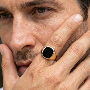 May include: A gold ring featuring a square, black onyx stone. The ring is shown close-up on a finger. The ring has a polished finish and a classic design.