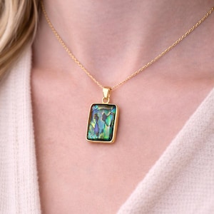 Puede incluir: Un collar dorado con un colgante rectangular con una incrustación iridiscente de concha de abulón. La concha muestra tonos de verde, azul y morado. El colgante está enmarcado en oro y cuelga de una delicada cadena dorada.