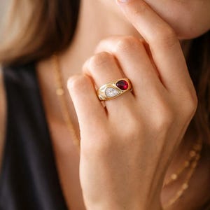 May include: A gold ring with a red gemstone and a white gemstone. The ring is on a finger. A gold hoop earring is visible in the background. The ring is a piece of jewelry.