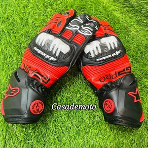 Könnte beinhalten: Ein Paar rot-schwarze Motorradhandschuhe mit weißen Akzenten, mit der Marke "Alpinestars" und dem Text "GP PRO". Die Handschuhe liegen auf grünem Gras, mit dem Text "Casademoto" sichtbar.