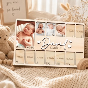 Könnte beinhalten: Ein rechteckiges hölzernes Meilenstein-Fotoboard für Babys mit einem großen Porträtfach, elf kleineren Fächern für die Monate 1 bis 12 und dem Namen David Darwin. Das Board enthält das Datum 01.01.2025 sowie dekorative Herz-Ausschnitte.