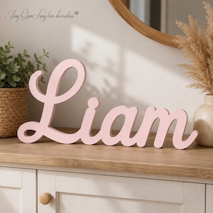 Könnte beinhalten: Ein hellrosa Holzschild mit dem Namen Liam in einer geschwungenen Schreibschrift. Die Buchstaben sind zu einem einzigen Stück verbunden und haben eine glatte, matte Oberfläche. Das Schild steht aufrecht auf einer ebenen Fläche.