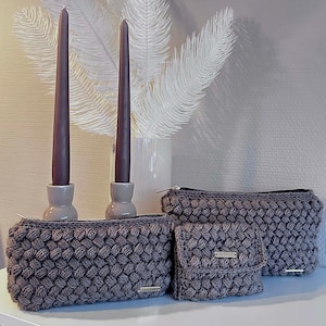 May include: A collection of gray crocheted bags and a small wallet with a textured, raised pattern. The bags vary in size, with a rectangular shape and a zipper closure. The wallet has a flap closure. Two purple candles in gray holders and a white vase with decorative feathers are in the background.