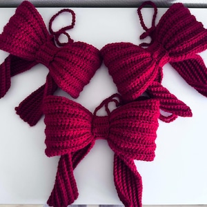 May include: Four handmade, crocheted bows in a deep red color. Each bow features a looped top for hanging and long, flowing tails. The bows are arranged on a white surface, showcasing their textured, ribbed design.