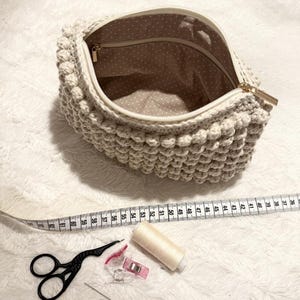 May include: A cream-colored, crocheted pouch with a textured, bobble stitch design. The interior features a dotted fabric lining. A gold zipper and a collection of sewing tools, including scissors, a measuring tape, and thread, are also visible.