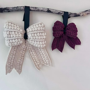 May include: Two handmade, textured bows hanging from a branch. One bow is a light beige color, and the other is a deep burgundy. Both bows are attached to black ribbons. The bows have a woven design and are likely made of yarn or fabric.