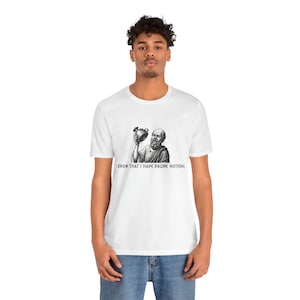 Puede incluir: Camiseta blanca con una ilustración en blanco y negro de un filósofo sosteniendo un recipiente. El texto "I KNOW THAT I HAVE DRUNK NOTHING" está impreso debajo de la imagen. La camiseta es de un material suave.