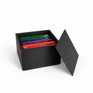 Könnte beinhalten: Eine schwarze Box mit leicht geöffnetem Deckel, die eine Reihe farbiger, rechteckiger Objekte im Inneren zeigt. Die Objekte sind in Grün-, Blau- und Rottönen gehalten. Die Box befindet sich auf weißem Hintergrund.