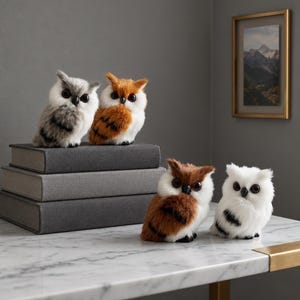 May include: Four small, plush owl figurines with soft, faux fur textures. Two owls are perched on a stack of three grey books, while two others sit on a white marble surface. The owls feature black beaks and large, dark, round eyes.