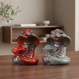 May include: Two ceramic cobra figurines. One is red and black, the other is grey and teal. Both have a smiling expression and a red tongue. The cobra figurines are decorative and likely for home decor.