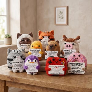 Op de afbeelding: Een verzameling gehaakte knuffels, waaronder een kat, een eend, een pinguïn, een capibara, een koe, een gamecontroller en een donut. Elk heeft een positieve boodschap op een klein bordje. De speeltjes zijn in verschillende kleuren en stijlen.