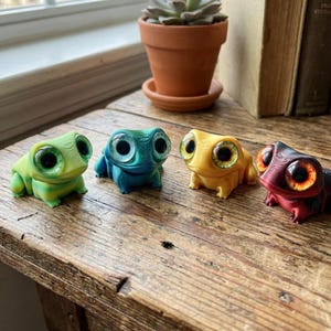 May include: Four brightly coloured frog ornaments in green, blue, yellow, and red, each with large eyes. They are arranged on a wooden surface, with a potted succulent and books in the background. Each frog has a unique colour and eye detail.