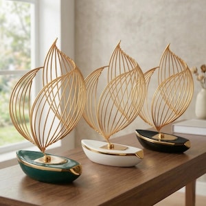 May include: Three decorative sculptures featuring gold wire leaf designs atop boat-shaped bases. The bases are in dark green, white, and black, each with gold trim. The sculptures are arranged on a wooden surface.
