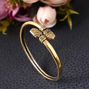 Könnte beinhalten: Ein goldfarbenes Metallarmband in offenem Design mit zwei detaillierten Tierkopf-Endstücken. Die Enden des Armreifs sind als stilisierte Köpfe mit gravierten Gesichtszügen und gerippten Bändern gestaltet, die eine durchgehende Kreisform bilden.