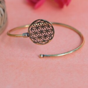 Può includere: Bracciale aperto color oro. Il bracciale presenta un design traforato circolare con un motivo geometrico. Il bracciale è in metallo e ha una piccola sfera all'estremità del bracciale. Il bracciale è su sfondo rosa.