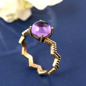 Pode incluir: Um anel dourado com uma grande pedra preciosa roxa redonda. A banda tem um design em zigue-zague de um lado e uma textura de contas do outro. O anel está sobre um fundo azul.