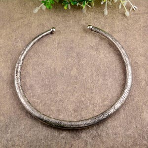 Puede incluir: Un collar tipo torque de metal plateado que presenta una banda circular continua con una abertura frontal. La superficie del collar está decorada con un intrincado patrón floral y geométrico en relieve, terminado con pequeños extremos redondeados.