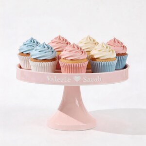 May include: A pink cake stand displays eight cupcakes with pastel-coloured frosting. The cupcakes have blue, pink, and yellow frosting. The cake stand has the names "Valerie" and "Sarah" with a heart symbol.