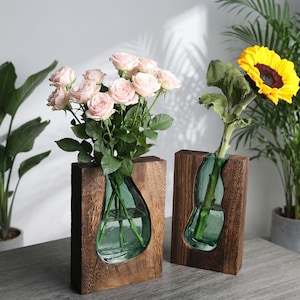 May include: Two wooden vases with a teardrop-shaped glass insert. One vase holds pink roses, the other a sunflower. The vases are made of dark wood and sit on a grey surface, with green plants in the background.