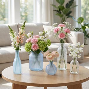 May include: A collection of fresh flowers in various glass vases on a round wooden table. The flowers include pink roses, carnations, and white sweet peas. The vases are in shades of blue and clear glass, creating a bright and airy display.
