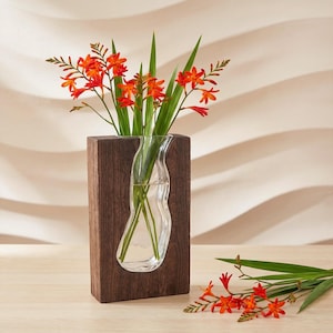 May include: A wooden vase with a clear glass insert, displaying orange flowers and green leaves. The vase is rectangular with a dark brown finish. The glass insert has a distinctive, curved shape. The flowers are a bright orange colour.