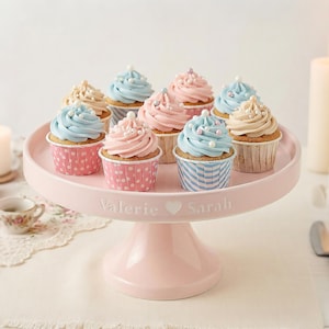 May include: A pink cake stand displays cupcakes with pastel-coloured frosting. The cupcakes feature blue, pink, and beige frosting, adorned with sprinkles. The stand is inscribed with "Valerie ♡ Sarah".
