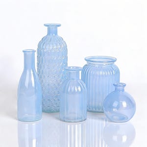 May include: A collection of five light blue glass vases of varying shapes and sizes. The vases include a tall, textured bottle, a slender bottle, a round vase, and two other vases with ribbed and patterned designs. The vases are arranged on a white surface.