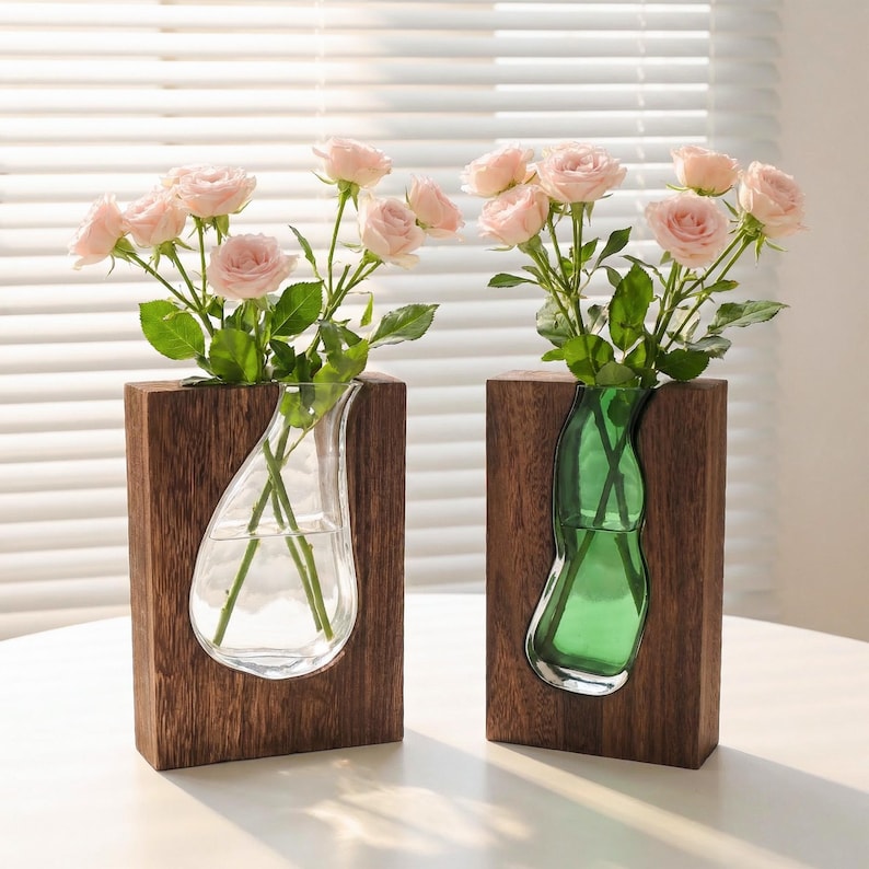 May include: Two wooden block vases with glass inserts, each holding pink roses. One vase has a clear glass insert, the other a green one. The vases sit on a white surface, with a window and blinds in the background.