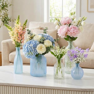 May include: A collection of fresh flowers in various glass vases on a white table. The arrangements include pink peonies, blue hydrangeas, and other colorful blooms. The vases are in shades of blue and clear glass, creating a bright and inviting display.