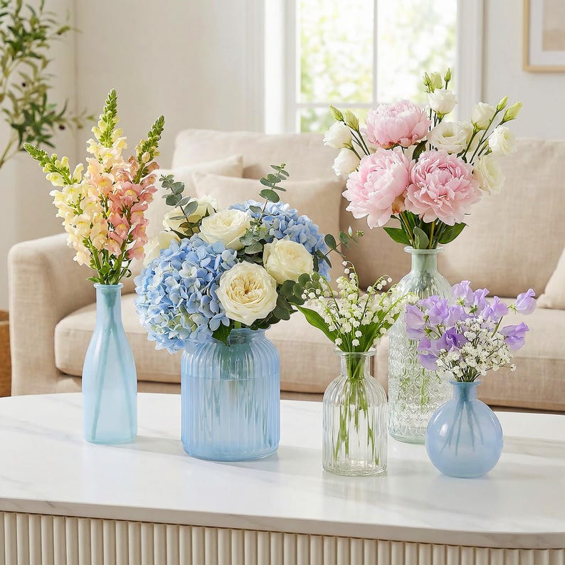 May include: A collection of fresh flowers in various glass vases on a white table. The arrangements include pink peonies, blue hydrangeas, and other colorful blooms. The vases are in shades of blue and clear glass, creating a bright and inviting display.