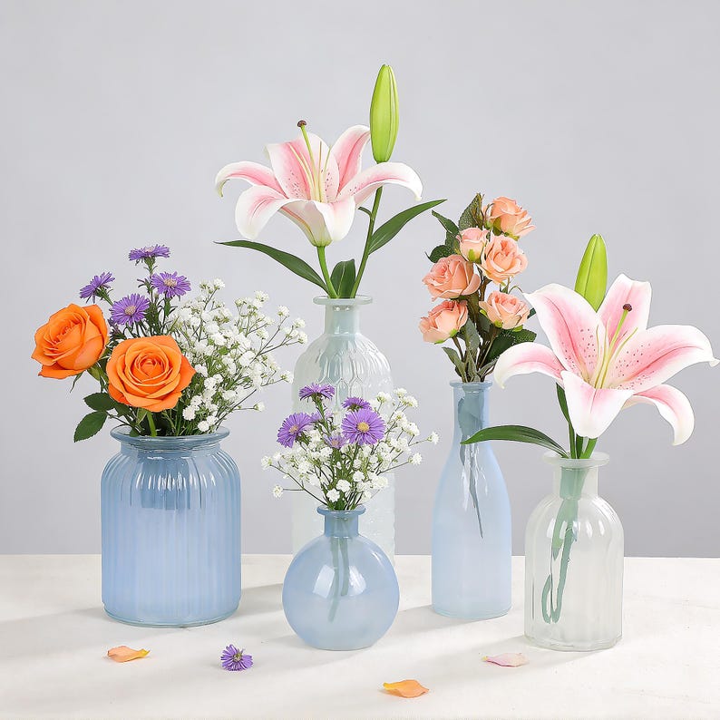 May include: A collection of glass vases in various shapes and sizes, each holding different types of flowers. The flowers include orange roses, pink lilies, and peach-colored roses. The vases are in shades of blue and clear.