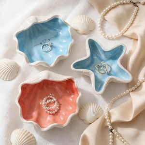 Pode incluir: Uma coleção de pratos de anel de cerâmica em forma de conchas e uma estrela do mar. Os pratos são pintados em tons de azul e coral, com detalhes brancos. Os pratos contêm anéis e são rodeados por conchas e um colar de pérolas.