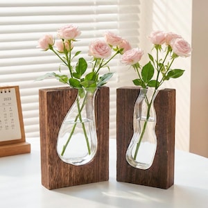 May include: Two wooden vases with clear glass inserts, each holding pink roses. The vases have a dark wood finish and a unique teardrop-shaped glass design. A 2023 calendar is visible in the background. A stylish home decor item.