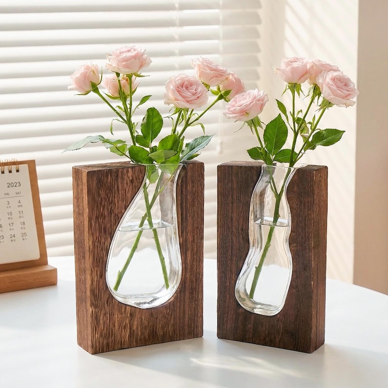 May include: Two wooden vases with clear glass inserts, each holding pink roses. The vases have a dark wood finish and a unique teardrop-shaped glass design. A 2023 calendar is visible in the background. A stylish home decor item.