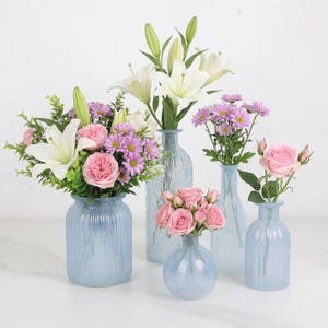 May include: An assortment of fresh flowers in various light blue glass vases. The flowers include white lilies, pink roses, and purple daisies. The vases are different shapes and sizes, creating a decorative display.