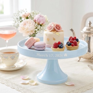 May include: A pale blue cake stand displays a small cake with rose decorations, macarons, and fruit tarts. The stand is inscribed with "March 17, 2026". A teacup, a glass of pink drink, and flowers are in the background.