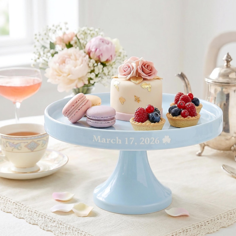 May include: A pale blue cake stand displays a small cake with rose decorations, macarons, and fruit tarts. The stand is inscribed with "March 17, 2026". A teacup, a glass of pink drink, and flowers are in the background.