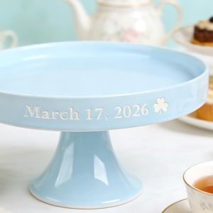 May include: A pale blue ceramic cake stand with a wide, flat top and a pedestal base. The date "March 17, 2026" is inscribed in white, along with a shamrock symbol. Ideal for displaying cakes and pastries at celebrations.