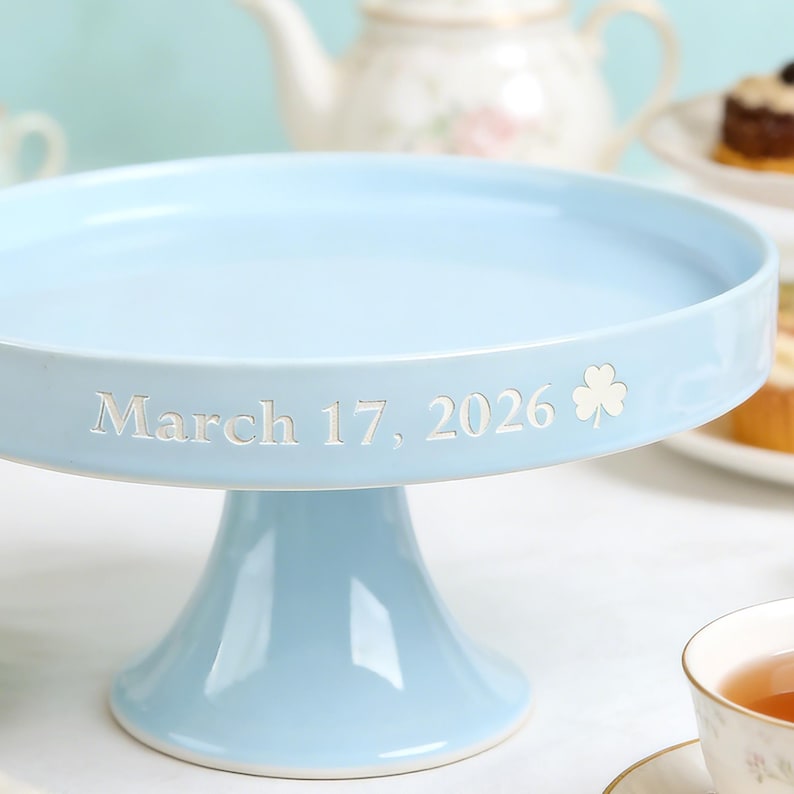 May include: A pale blue ceramic cake stand with a wide, flat top and a pedestal base. The date "March 17, 2026" is inscribed in white, along with a shamrock symbol. Ideal for displaying cakes and pastries at celebrations.