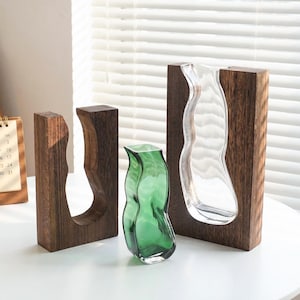 May include: Three decorative vases. One is clear glass, another is green glass, and the third is an open wooden frame. The vases have organic, flowing shapes. The wooden frames are dark brown and rectangular.