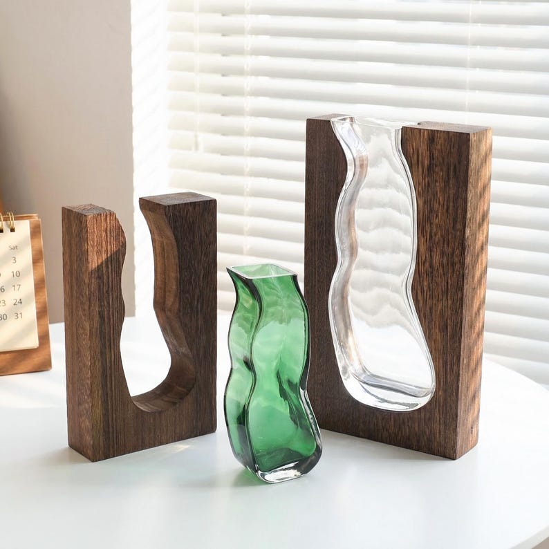 May include: Three decorative vases. One is clear glass, another is green glass, and the third is an open wooden frame. The vases have organic, flowing shapes. The wooden frames are dark brown and rectangular.