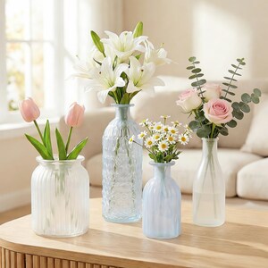 May include: A collection of glass vases with various floral arrangements. The vases are different shapes and sizes, with clear and frosted finishes. Flowers include white lilies, pink roses, daisies, and pink tulips.