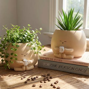 May include: Two beige ceramic planters shaped like figures, each holding a small mug. One contains a trailing green plant, the other a spiky succulent. The planters sit on a wooden surface with coffee beans.