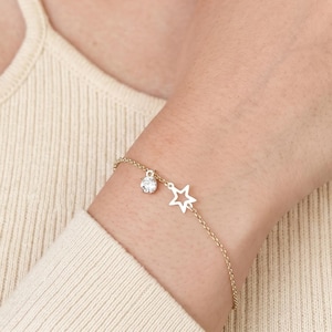 May include: A delicate gold-tone chain bracelet featuring a small, open-frame star charm and a single round, clear faceted crystal charm. The bracelet is secured with a thin metal chain, showcasing a minimalist design with two distinct charms.