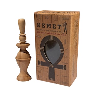 May include: A light brown wooden cosmetic applicator next to a cardboard box. The box features the word "KEMET" and an ankh symbol. Text on the box reads "This product is crafted specifically for you. 100% natural handmade body care products."